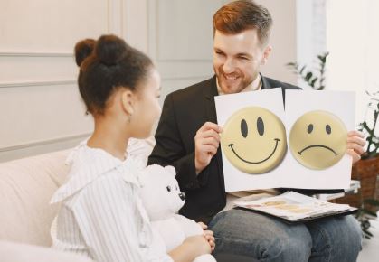 A man holding sad and happy emoji charts and showing it to a girl holding her teddy bear in hand