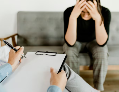 a person sitting holding their head - along with a lady who is holding the clipboard